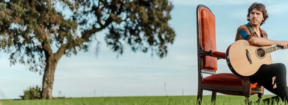 Alejandro Rivera sentado en el campo con una guitarra y un árbol al lado.